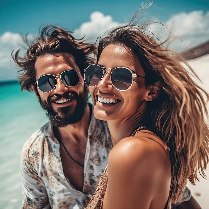 Couple heureux souriant sur une plage, homme brun bronzé avec lunettes et chemise ouverte, femme bronzée aux cheveux longs châtains clairs, portant des lunettes et un haut, symbole de bien-être et de vitalité.